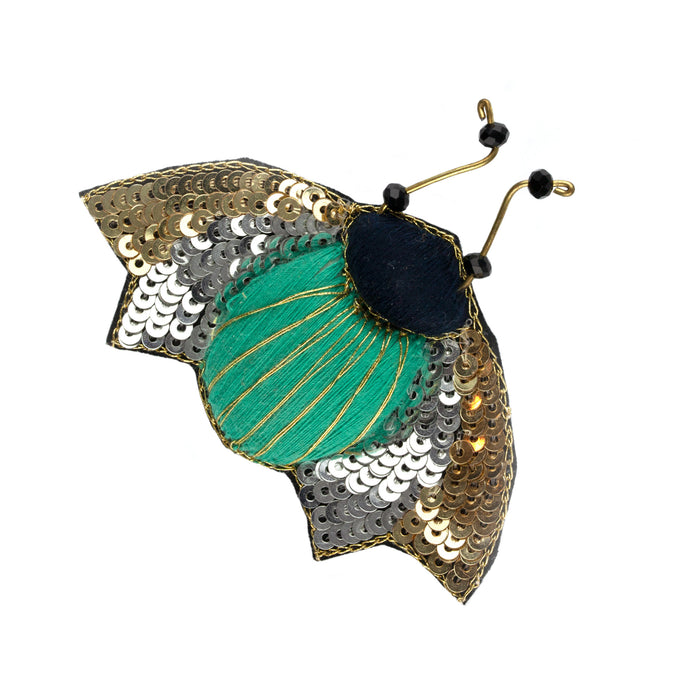 Brosch med en kropp av grönt tyg och guldtråd, vingar av silverpaljetter samt detaljer av metall och svarta pärlor.  Storlek 5,5 x 7,5 cm 