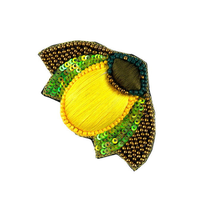 Brosch med en kropp av solgult tyg och gula pärlor, vingar av guldfärgade pärlor och gröna paljetter och ett huvud av brunt tyg och blå pärlor.  Storlek 4,5 x 7 cm.